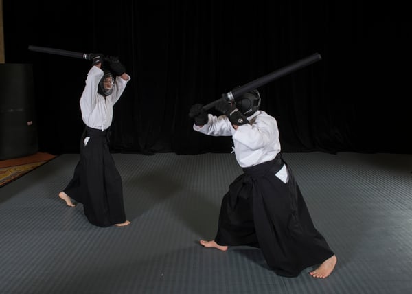 Two martial artists wearing face armor and helmets spar with foam katana.